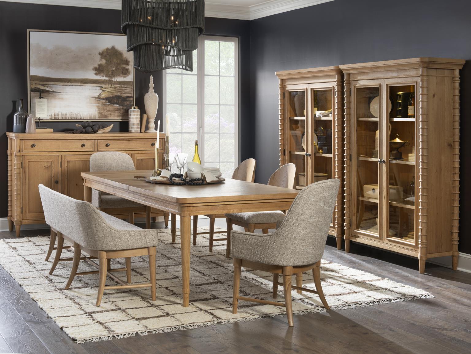 Light wood dining set with gray upholstered dining room benches and chairs, sideboard, and glass display cabinets in a dark blue room.