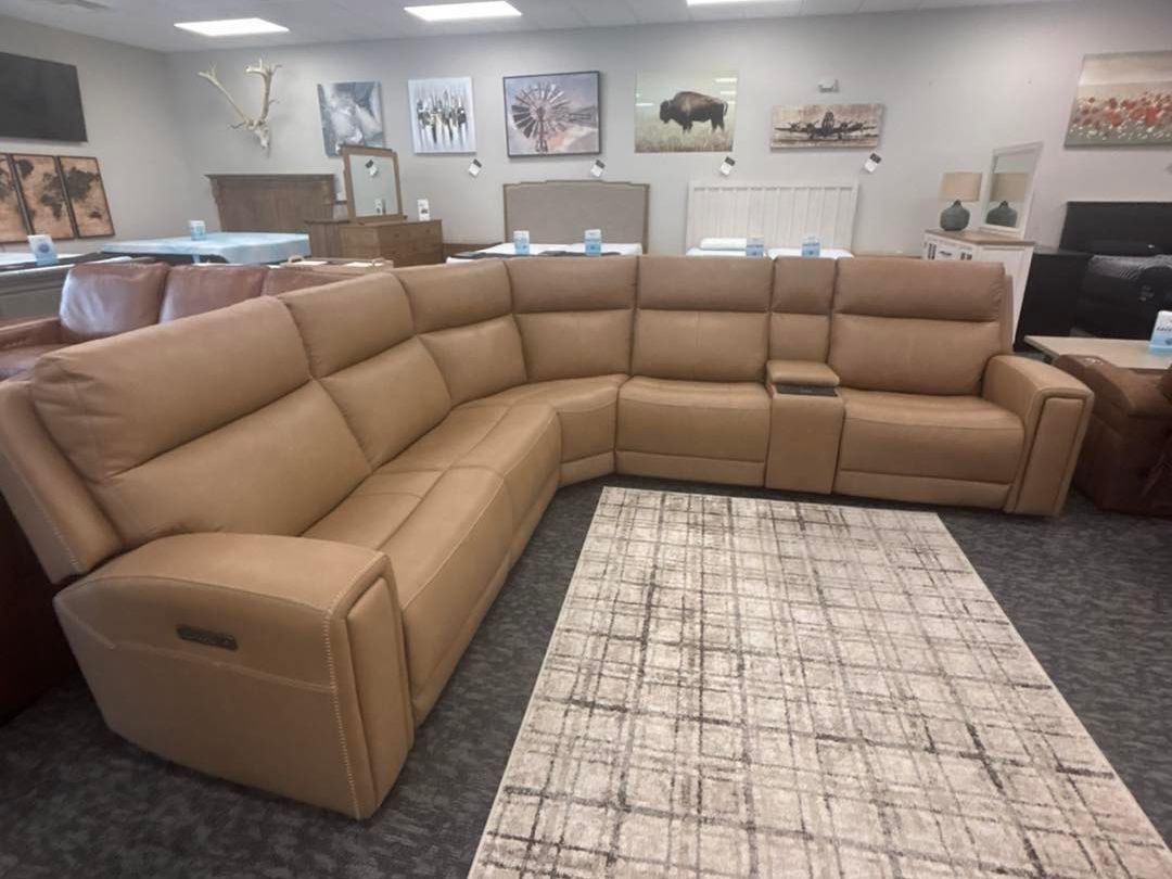 A spacious, light tan leather sectional sofa, at Bed'r Furniture & Mattress in Waycross, GA, arranged in a curved shape, placed in a showroom with several artwork pieces on the walls and a light-colored rug beneath it.