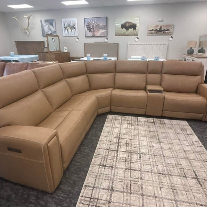 A spacious, light tan leather sectional sofa, at Bed'r Furniture & Mattress in Waycross, GA, arranged in a curved shape, placed in a showroom with several artwork pieces on the walls and a light-colored rug beneath it.